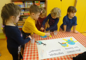 Piątka dzieci dekoruje plakat ekologiczny, naklejają elementy z kolorowego papieru.
